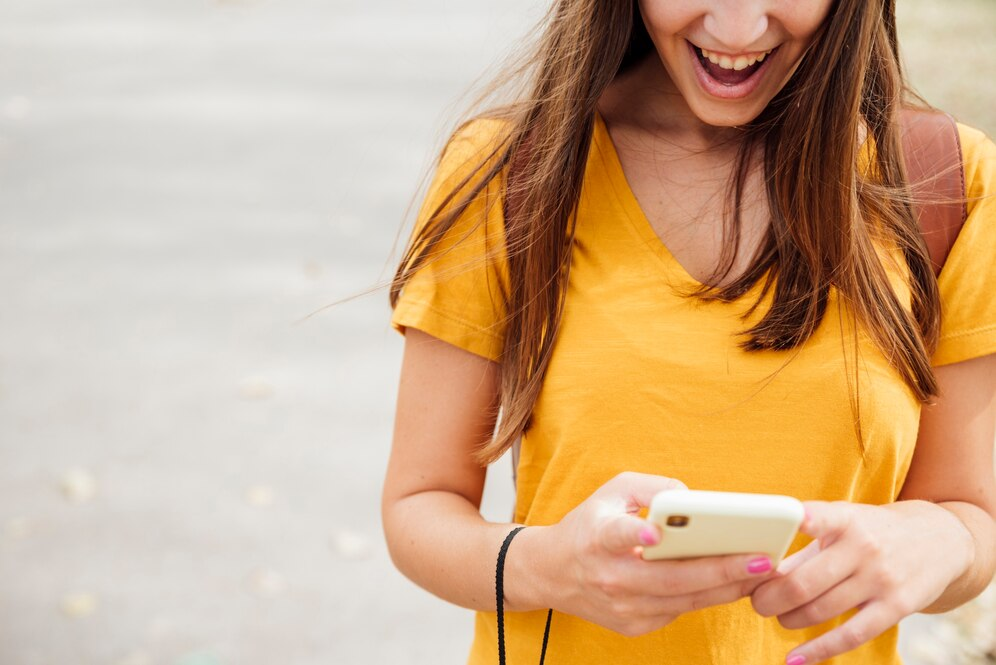 Mulher usando camiseta amarela olhando para o celular com uma expressão animada. Título: Surpresa Digita