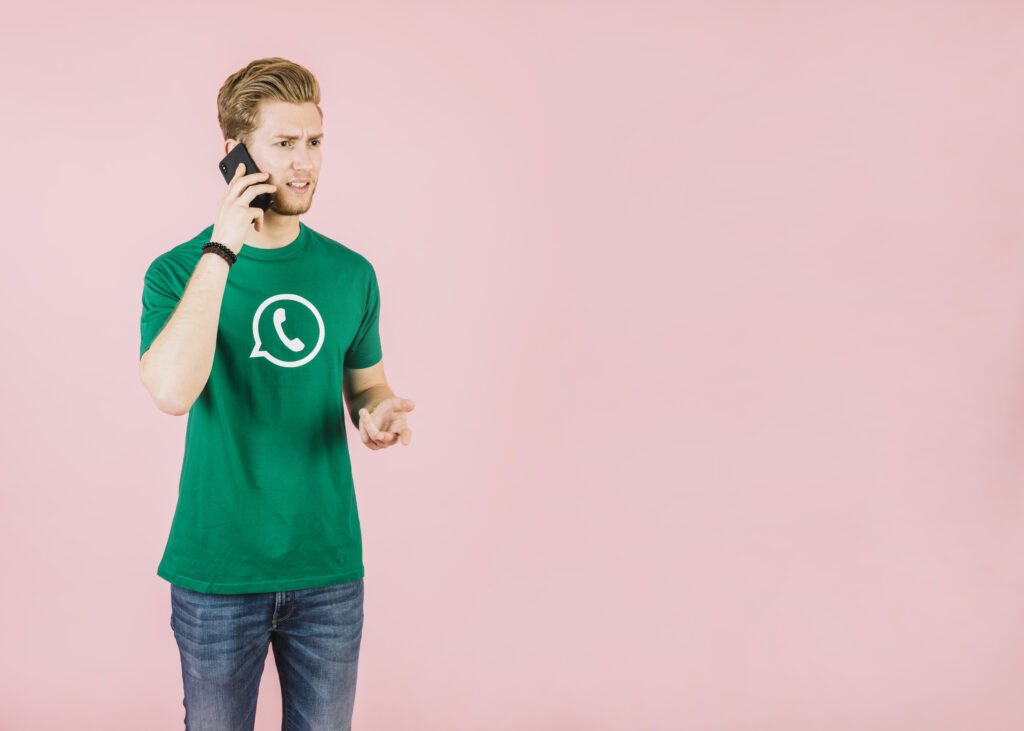 Homem jovem, usando uma camiseta verde com o símbolo do WhatsApp, segura um celular ao ouvido e expressa confusão, com fundo rosa liso.